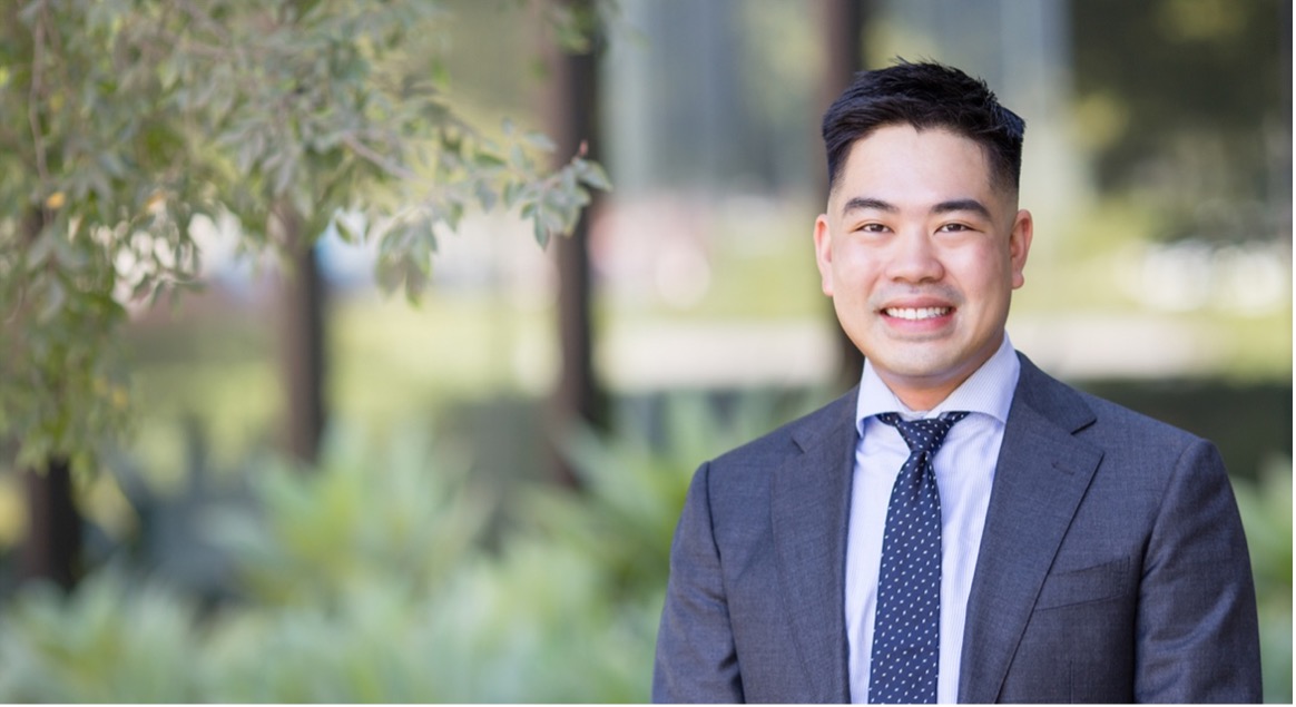 Smiling man with short black hair wearing a blue suit and tie standing in front of a leafy green background outside