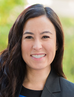 Smiling woman with long brown hair with greenery in the background.
