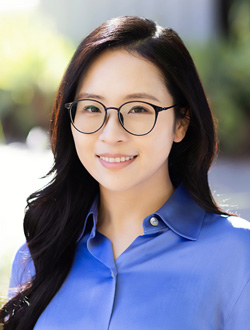Smiling woman with long dark hair, wearing glasses and a blue shirt with greenery in the background.