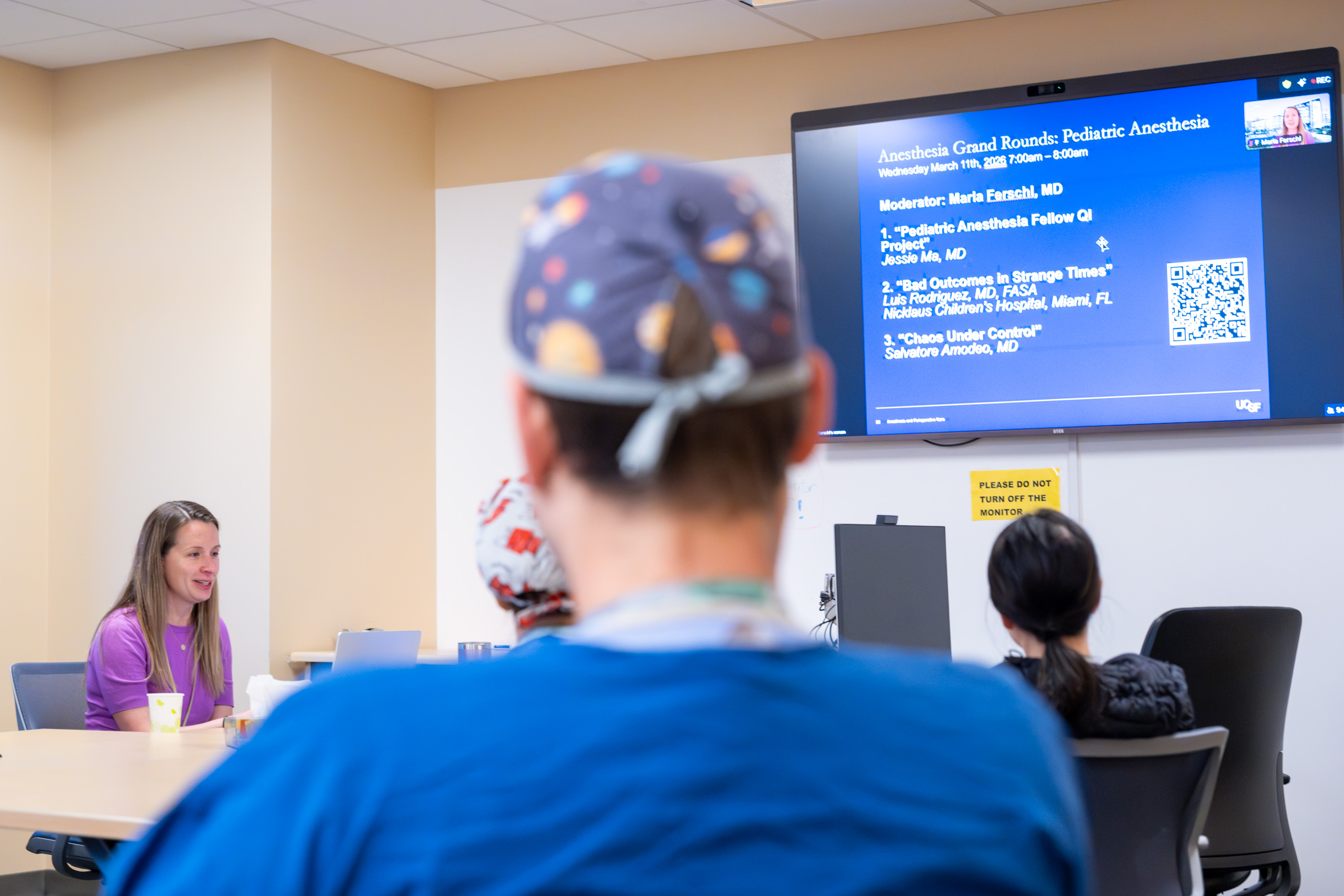 Dr. Ferschl seated at a conference table introducing the Grand Rounds with an audience looking on and a large display in the background showing the Zoom portion of the meeting.