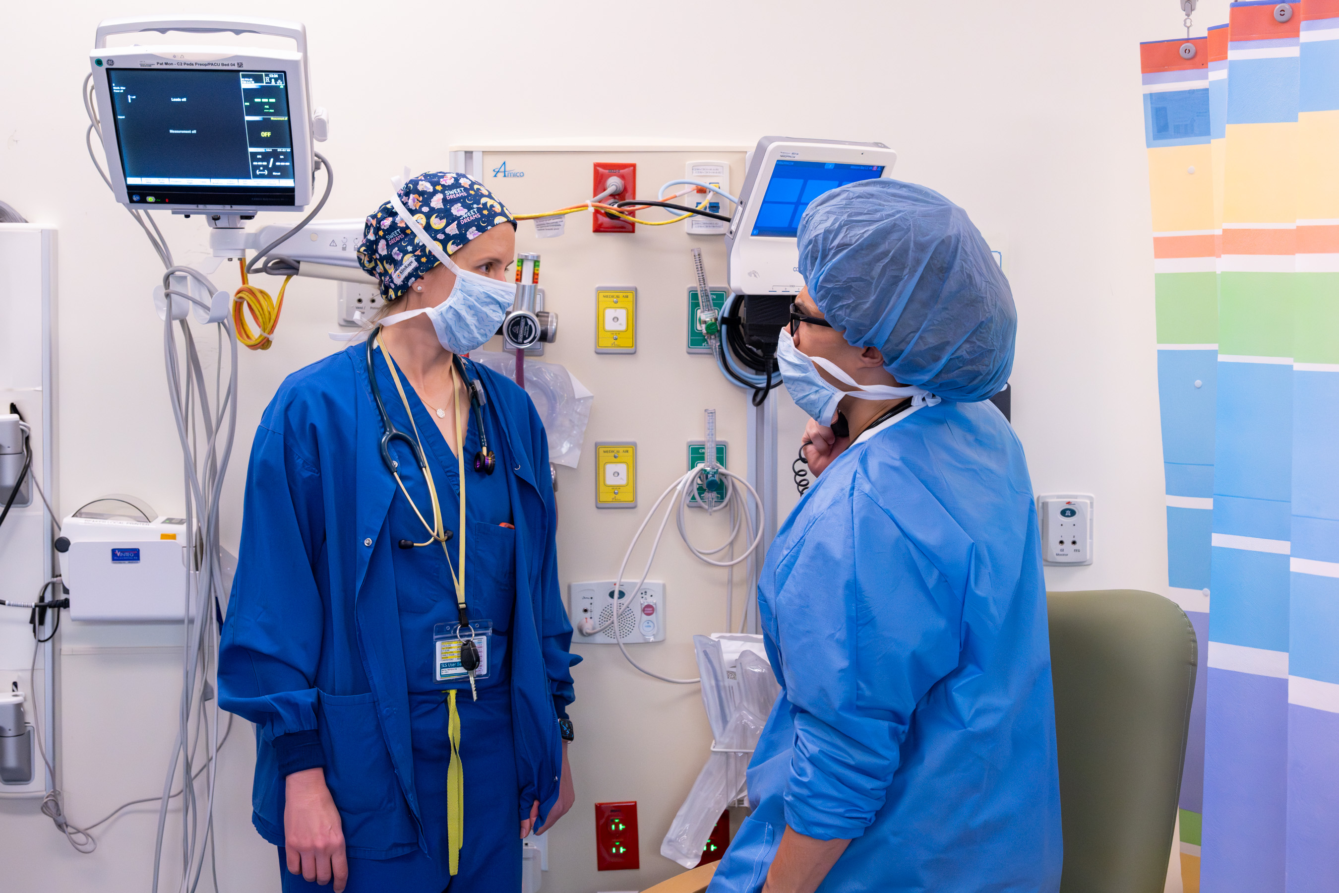 Two people wearing blue scrubs, masks and head coverings talk in a clinical setting.