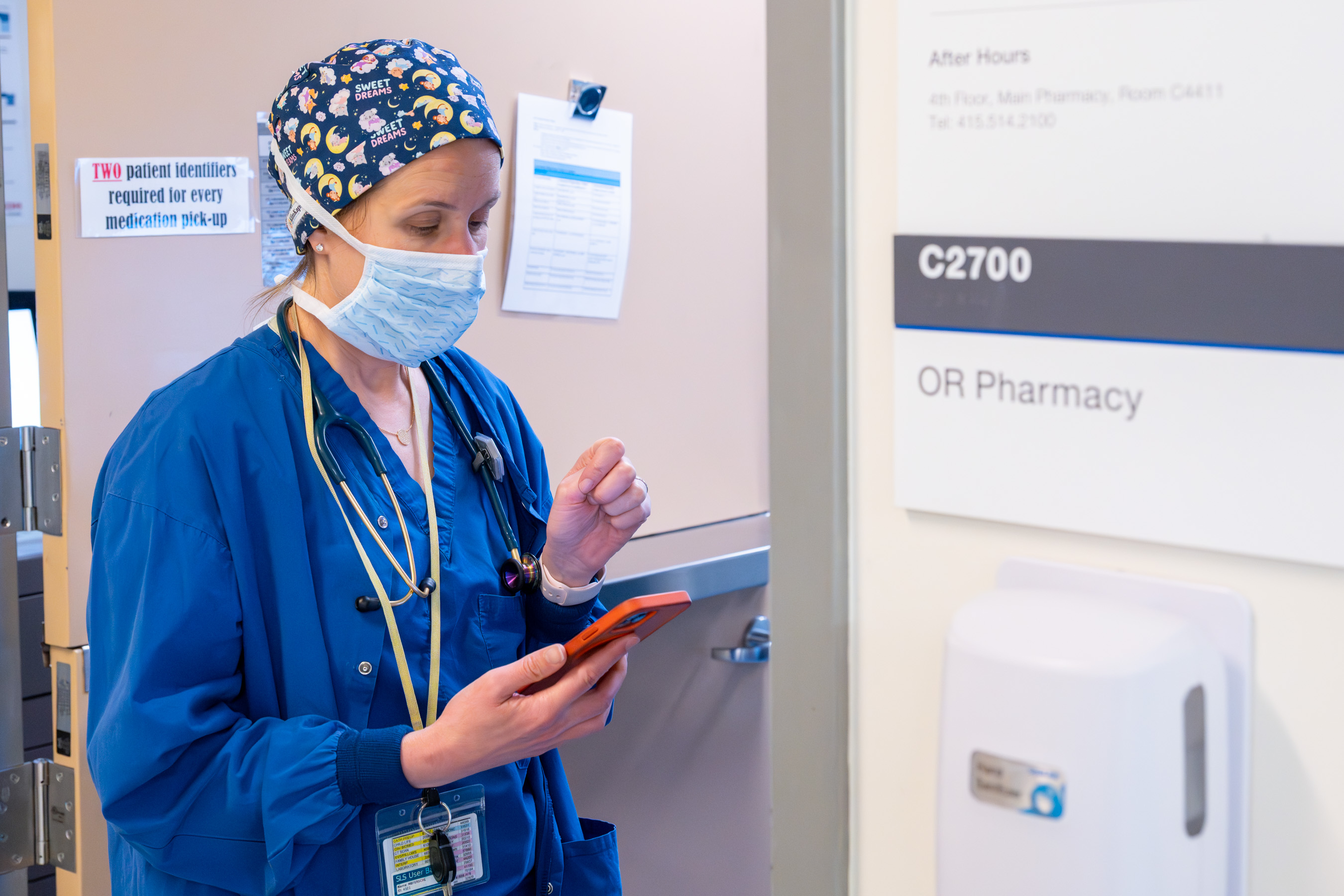 Dr. Ferschl wearing scrubs, a mask and head covering at the OR Pharmacy checking with the pharmacist and referring to her phone.