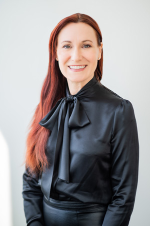 Carroll Schreibman smiling, and wearing a black shirt with long red hair and a white background.