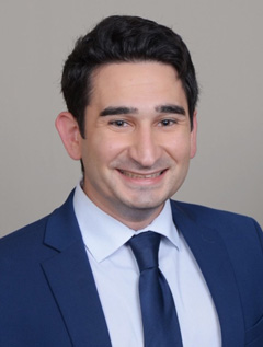 Smiling person wearing a blue suit and tie with short black hair.