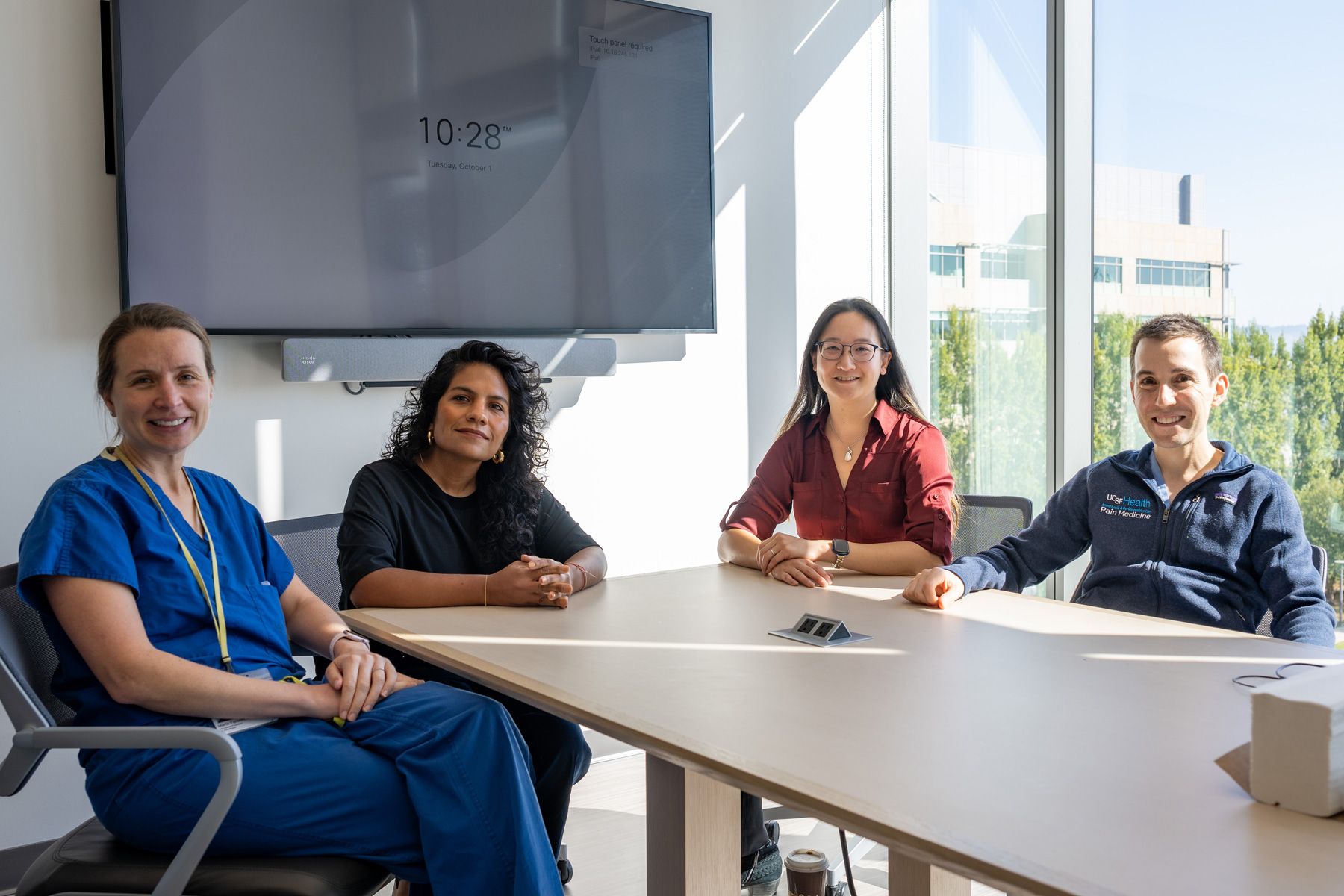 Four doctors seated at a conference table with floor to ceiling windows overlooking trees and a building in the distance