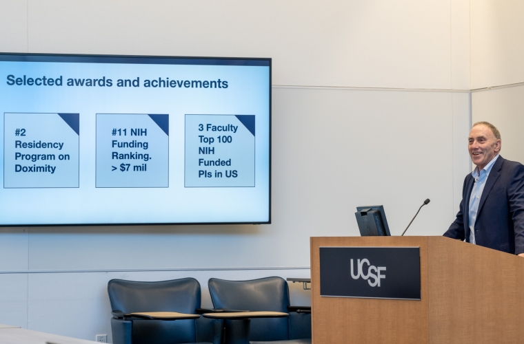 Smiling man in a navy blazer delivers remarks from a podium with a screen in the background