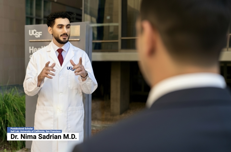 Man in a white UCSF lab coat standing outside delivering remarks to another person.