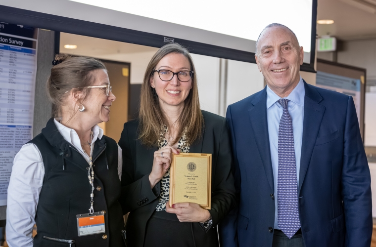 Three people smiling and standing together, the one in the middle holds a plaque.