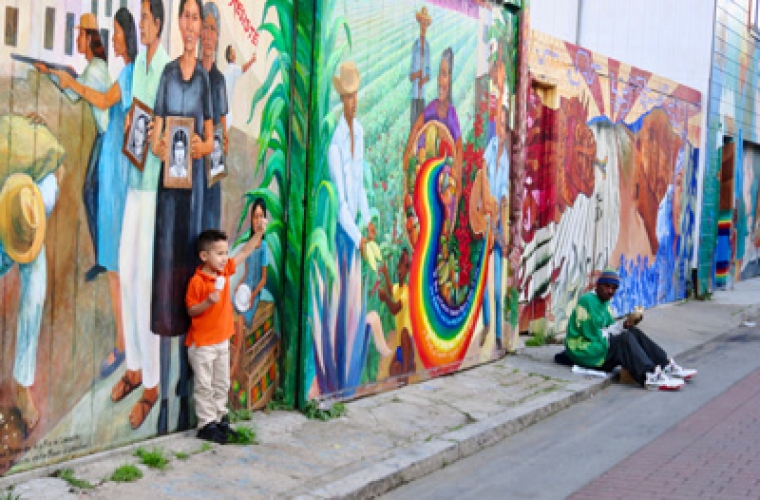 A young boy in a red shirt standing and waving in front of a large colorful mural.