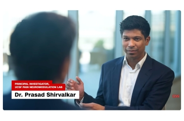 Dr. Prasad Shirvalkar, Principal Investigator, UCSF Pain Neuromodulation Lab, wearing a white collared shirt and dark blue jacket speaks with another person in front of large windows.