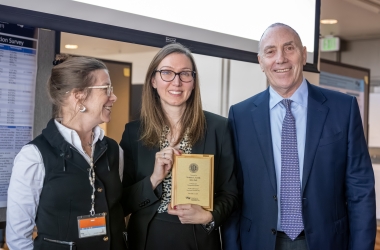 Three people smiling and standing together, the one in the middle holds a plaque.