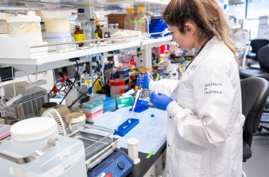 A woman with long light brown hair standing at a bench in a lab setting using a pipette wearing a white lab coat that is embroidered with the words, "University of California."