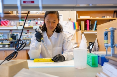A person with long black hair wearing a lab coat sits at a lab bench using a pipette to fill a sample vial in a lab environment.