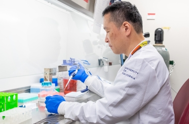 Person at a lab bench with a hood wearing blue gloves and a white lab coat using a pipette.
