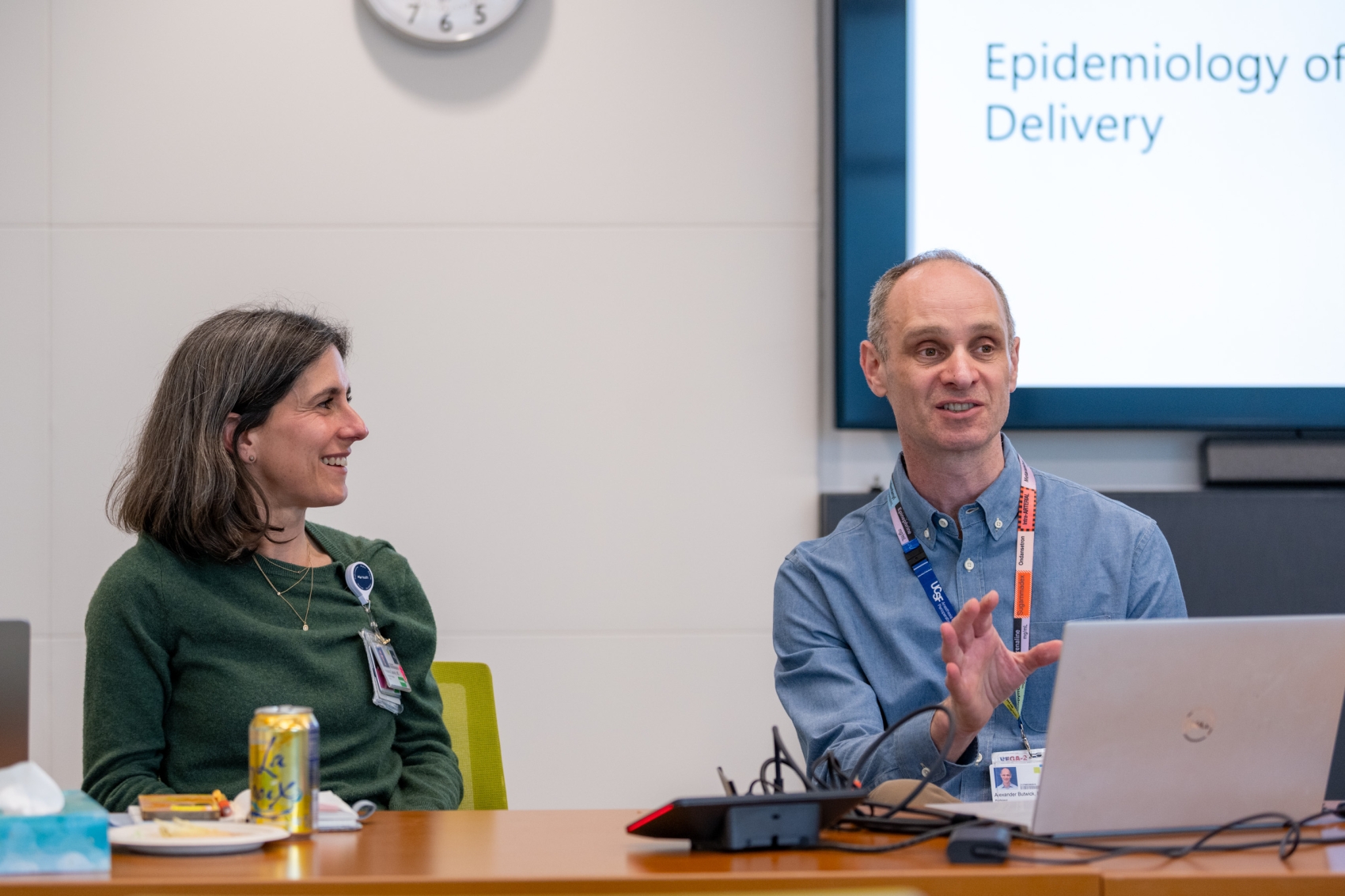 Two people sitting at a table, the person on the right addressing an audience while the person on the smiles and looks on.