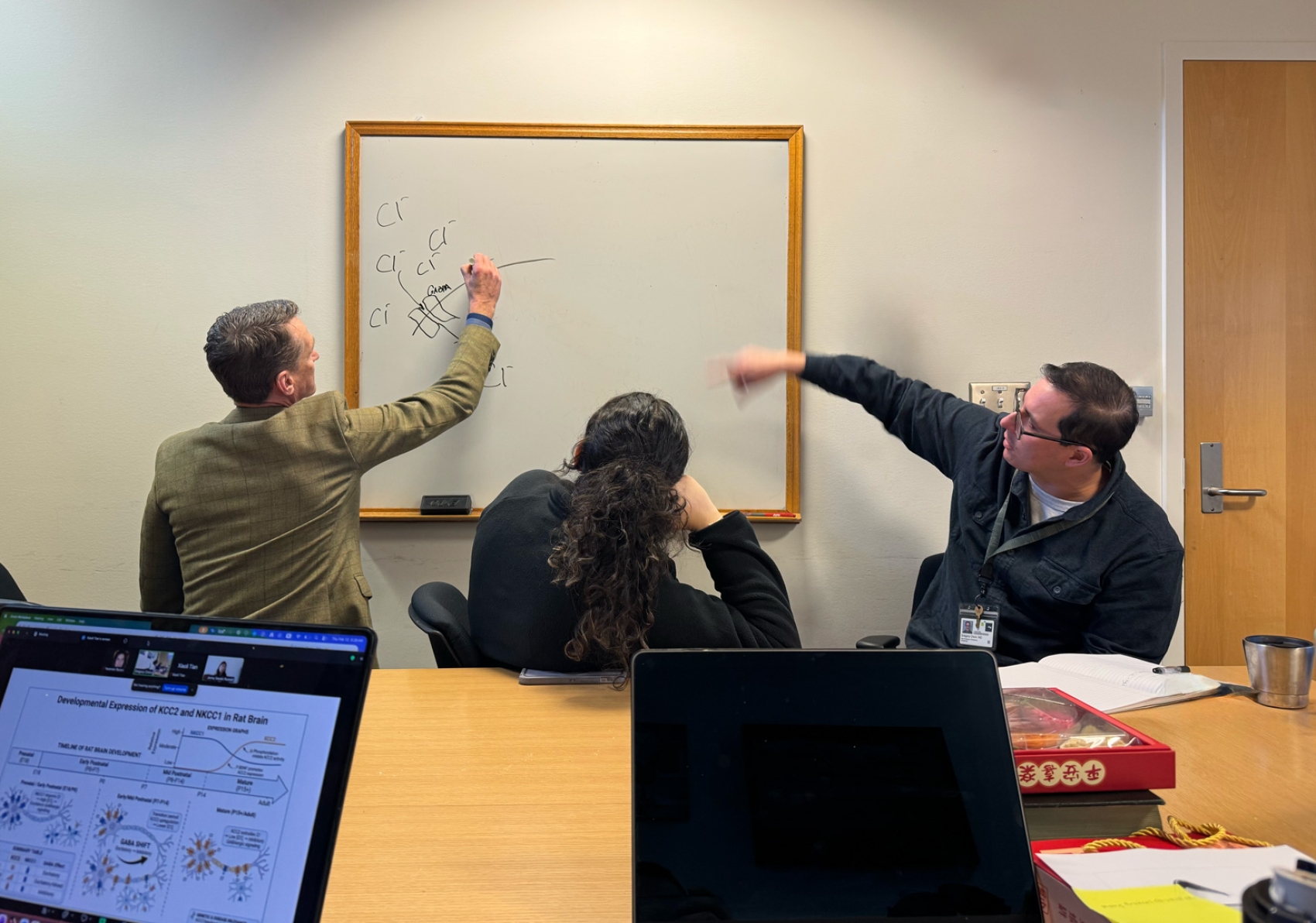 Three people around a white board, one person writing on the board, one pointing to the board and one looking on.