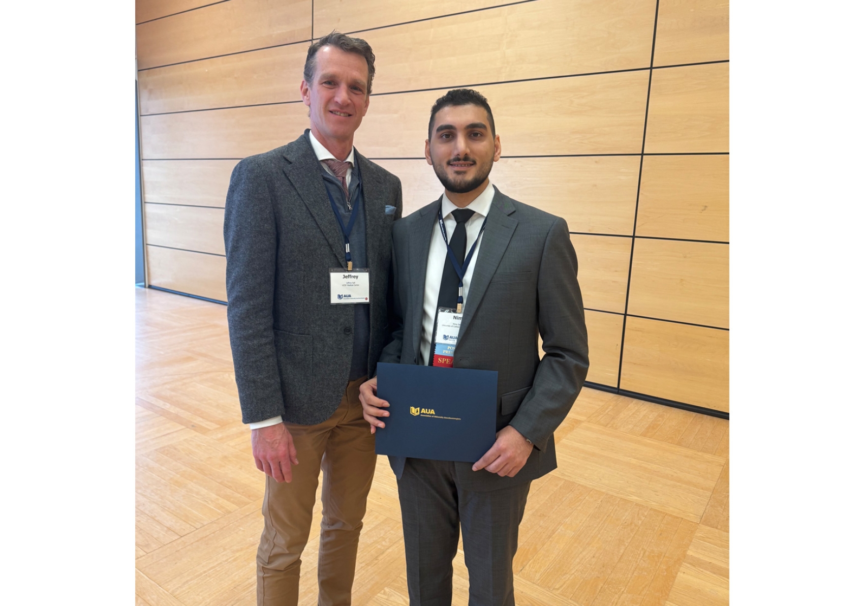 Two people in suits and ties standing together, one holding a certificate folder.