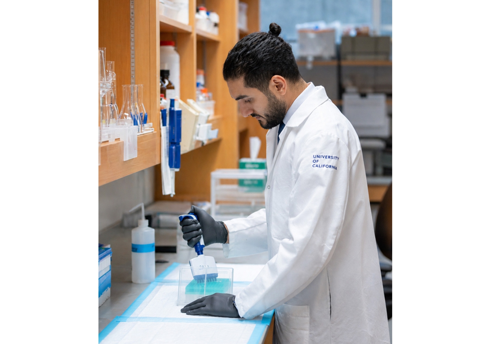 Person wearing a white lab coat and black gloves standing at a lab bench using a multichannel pipette.