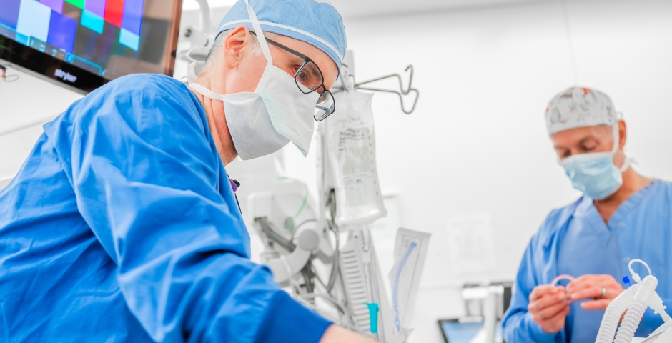 Two men wearing blue scrubs, mask, and head coverings in an operating room setting.