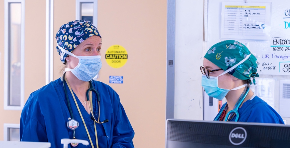 Two doctors wearing scrubs, masks and head coverings standing and talking in a clinical setting.