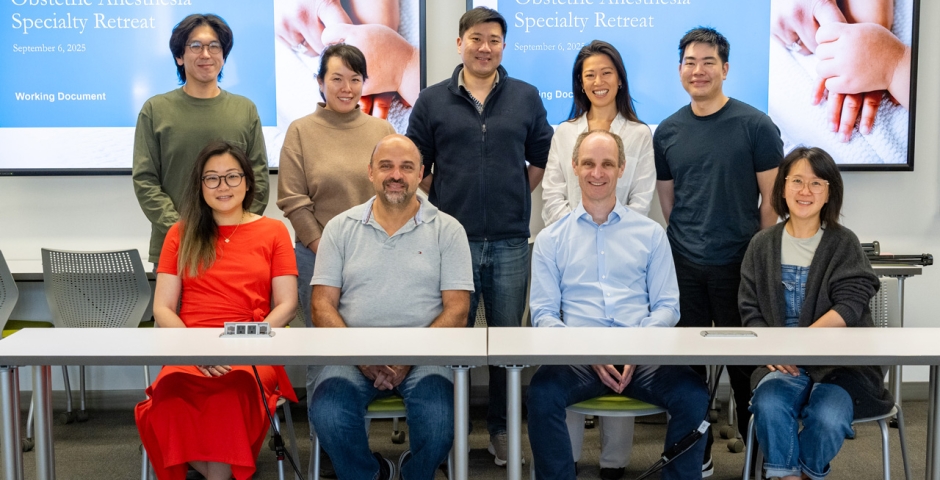 A group of nine doctors posing together in front of a large digital screen that reads "Obstetric Anesthesia Specialty Retreat."