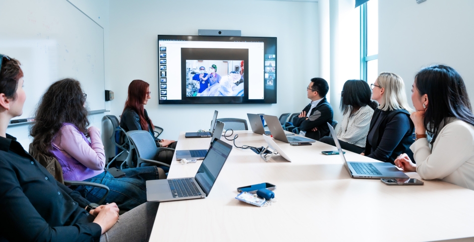 A group of professionals seated at a large conference table with laptops looking at a large display in the background showing an image of medical professionals in a clinical setting.