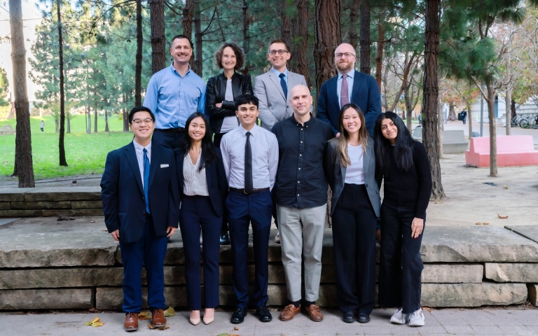 Group of people in formal business attire sanding in two rows together outside with trees in the background