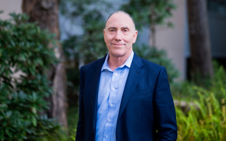 Man wearing a navy blazer over a light blue button down shirt standing outside with trees and shrubs in the background.