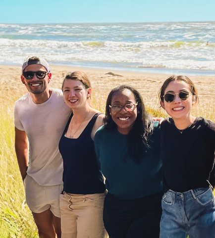Group of four people standing arm-in-arm on a beach with the ocean in the background.