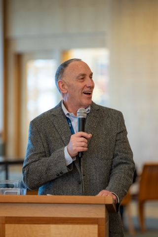 A man standing at a podium speaking into a microphone while smiling and looking to his left.