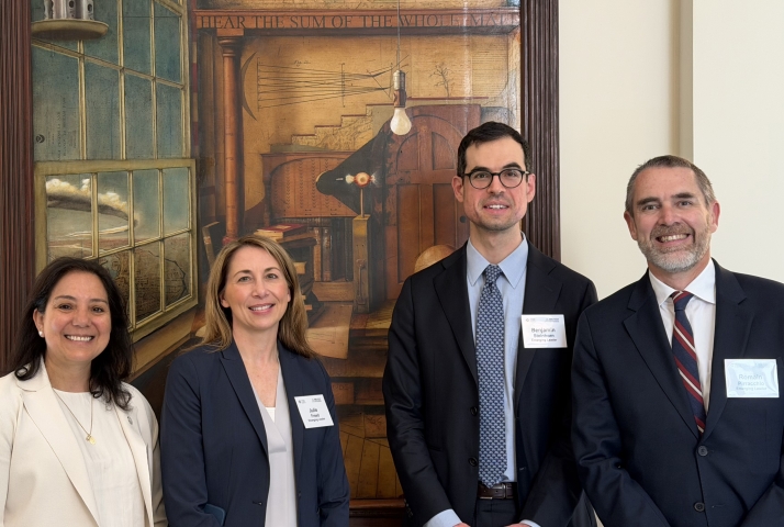 Four people wearing suits with nametags standing together and smiling in front of a large painting..