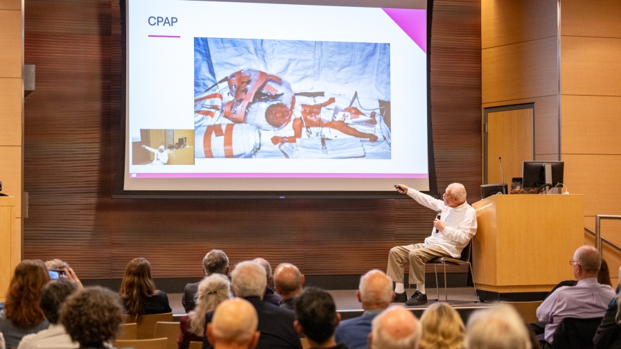 Man in shite long sleeve shirt giving a presentation in front of an audience while pointing at a slide of an early CPAP device