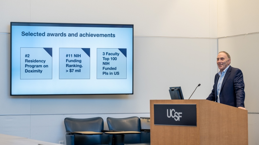 Smiling man in a navy blazer delivers remarks from a podium with a screen in the background