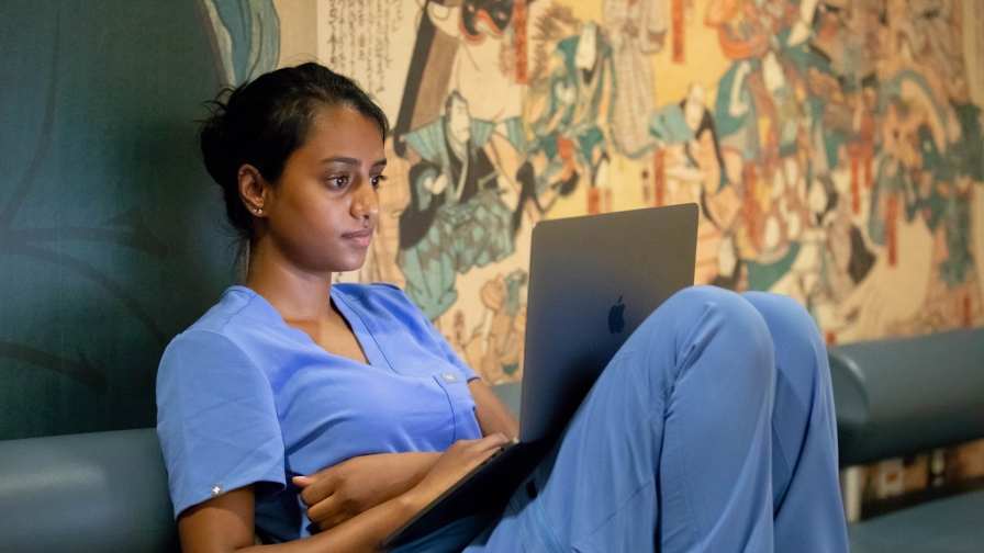 Person with long dark hair pulled back, wearing blue scrubs and intently studying the screen of the laptop