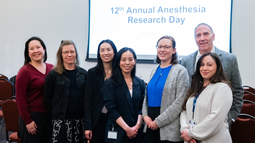 A group of seven people standing together in a conference space with a large projector screen which reads "12th Annual Anesthesia Research Day"