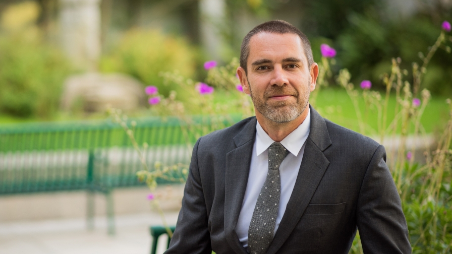 Man with short hair and a grey and brown beard wearing a suit and tie sitting on bench with flowers in the background.