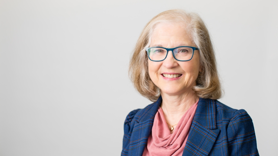 Woman with blue framed glasses and should length blond hair smiling, wearing a pick sweater under a navy blazer, with a gray background.