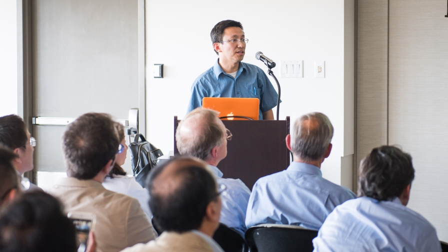 Man with short dark hair, wearing glasses, standing at a podium in with a microphone delivering remarks to an audience in the foreground.