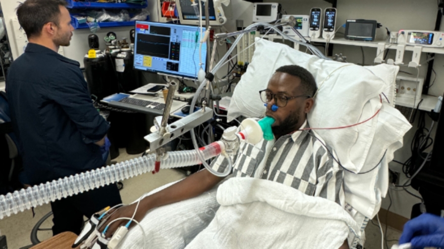 Man with short black hair and glasses laying in a hospital bed with a respiration tube and pulse oximeters connected to each finger on his right hand while another man monitors a computer display next to him.