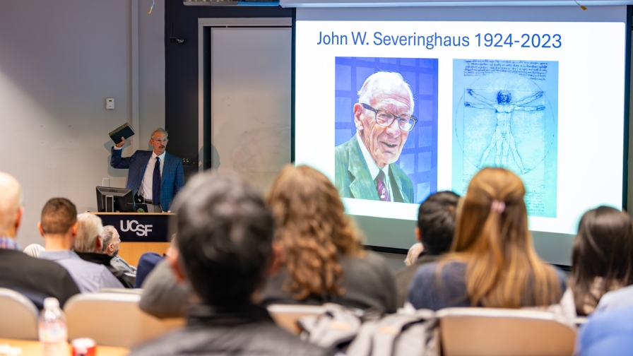 An audience watching a man at a podium speaking while holding a book with a slide projected in the background reading "John W Servinghaus 1924-2023".