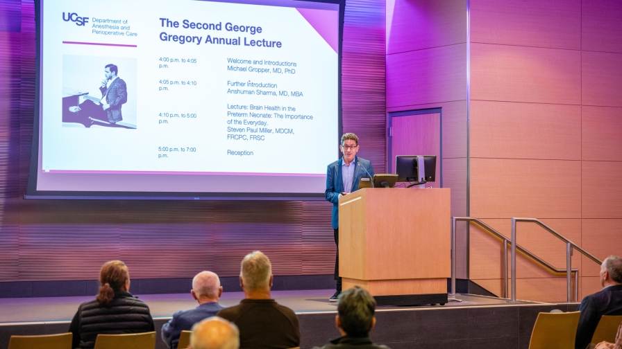 Man standing at a podium on a stage delivering remarks with a large projected slide behind him.