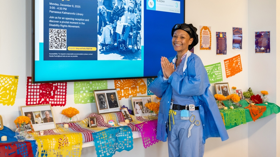 Smiling woman in scrubs standing in front of a colorful ofrenda featuring framed photos and flowers.