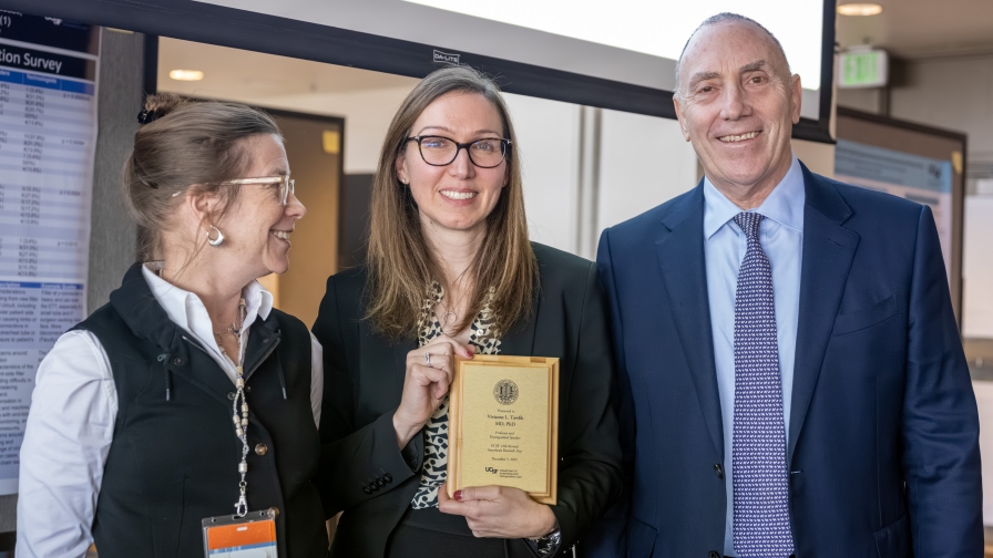 Three people smiling and standing together, the one in the middle holds a plaque.