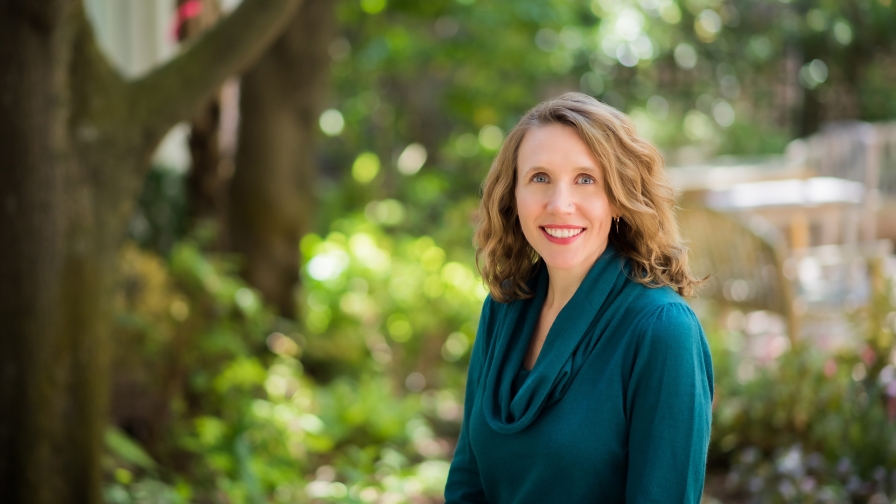 A smiling woman with long wavy light brown hair standing in a garden wearing a blue-green sweater.