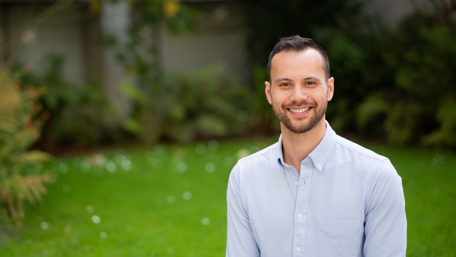 A smiling man with a short brown beard and short brown hair wearing a light blue shirt with grass and greenery in the background.