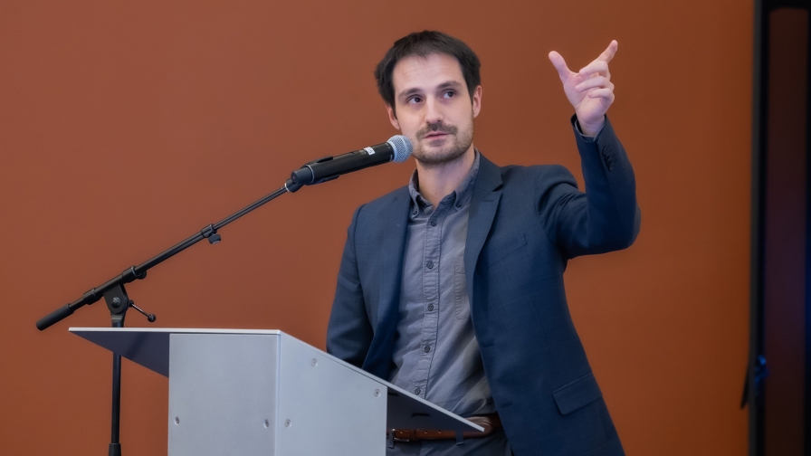 Person with short dark hair wearing a gray shirt and dark gray jacket standing at a lectern and speaking into a microphone while gesturing.