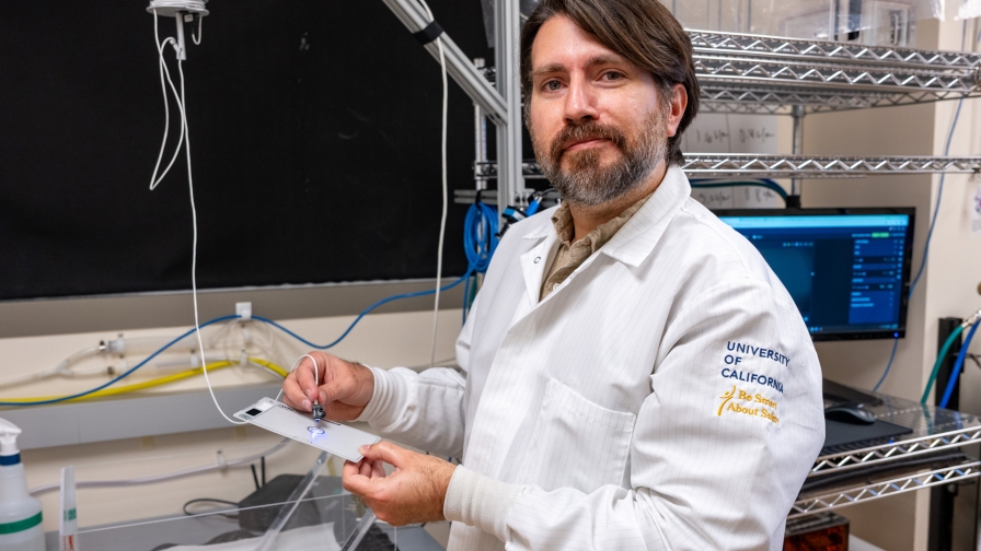 Person with brown hair and a beard wearing a white lab coat standing in a lab setting holding a light sensor.