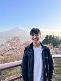 Man in white shirt and black jacket stands on balcony in front of mountain.