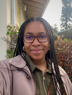 Woman smiling with long braids and glasses, in the background there is a tree and part of a building