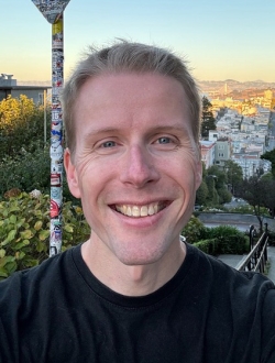 Man with short blond hair, smiling, and wearing a black tee-shirt, with a view of San Francisco in the background